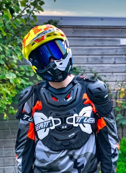 The fox stands with his head slightly tilted to the left in his full MX outfit in a garden with a shed and some plants visible in the background. He looks at the camera and is in focus while the background is slightly blurred. The full MX set of the fox consists of a grey camo-patterned FOX MX jersey with orange text highlights, a black-white Leatt MX chest protector, a yellow-orange MX helmet and borderless dark-tinted MX goggles.