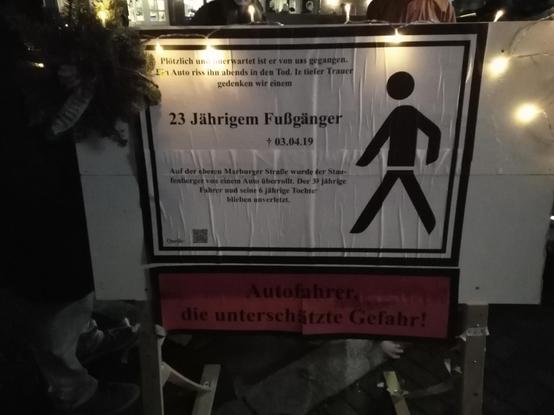 Trauerschild auf Ladezone: "Plötzlich und unerwartet ist er von uns gegangen. Ein Auto riss ihn abends in den Tod. In tiefer Trauer gedenken wir einem

[hervorgehoben] 23 Jährigem Fußgänger
[Kreuzsymbol] 03.04.19

Auf der oberen Marburger Straße wurde der Staufenberger von einem Auto überrollt. Der 39-Jährige Fahrer und seine 6 Jährige Tochter blieben unverletzt."
Rechtsbündig ist ein Fußgängysymbol abgebildet.
Unterhalb des Schildes steht rot unterlegt, im Stil einer Tabakwarenwarnung: "Autofahrer, die unterschätzte Gefahr!"

Links oben am Schild ist ein Trauergesteck angebracht und oberhalb am Schild verläuft eine Lichterkette .