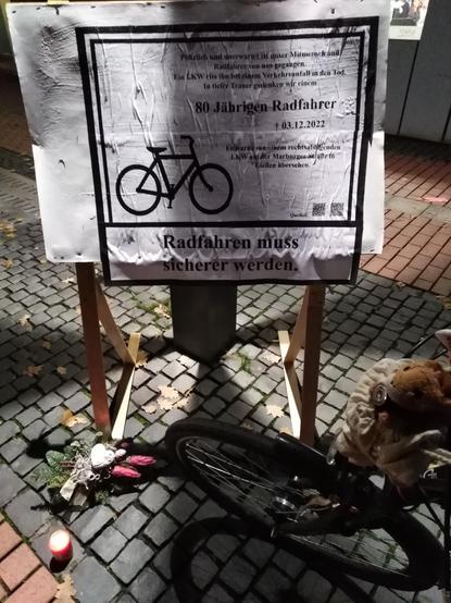 Gedenkschild bei Nacht auf Fußgängyweg. Am Fuß des Stehschildes liegt ein Trauergesteck davor eine Grabkerze.
Auf dem Schild ist rechtsbündig ein Text geschrieben in Form einer Traueranzeige: 
"Plötzlich und unerwartet ist unser Mitmensch und Radfahrer von uns gegangen. Ein LKW riss ihn bei einem Verkehrsunfall in den Tod. In tiefer Trauer gedenken wir einem 80 Jährigen Radfahrer. Kreuzsymbol (gestorben) 3.12.2022
Er wurde von einem rechtsabbiegenden LKW auf der Marburgerstr. in Gießen übersehen."
Linksbündig unten ist ein Fahrradsymbol abgebildet.
Unterhalb ist noch ein weiterer Text in Form einer Zigarettenschachtelwarnung angebracht: "Radfahren muss sicherer werden." 

Puschel, das kleinste Eichhörnchen sitzt im Auskucksackerl vorne am Radlenker rechts neben dem Schild.