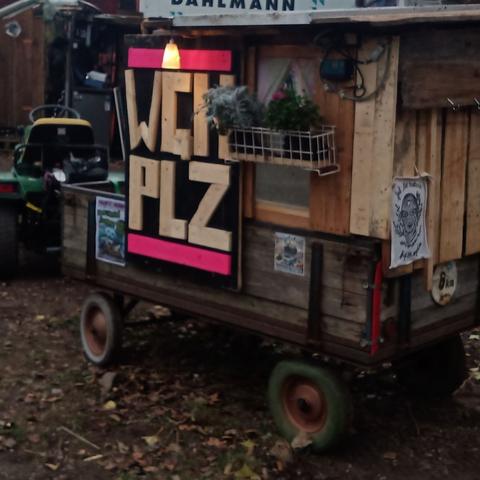 Zu srhen ist einer der Demo Wagen. Es ist ein kleiner Anhänger an einem RasenmäherTraktor. Er hat einen aufbau aus holz bekommen, vor einem fensterchen mit gardinen hängen blumentöpfe. Es hängen ein paar plakate daran und ein schild, auf dem WGN PLZ steht