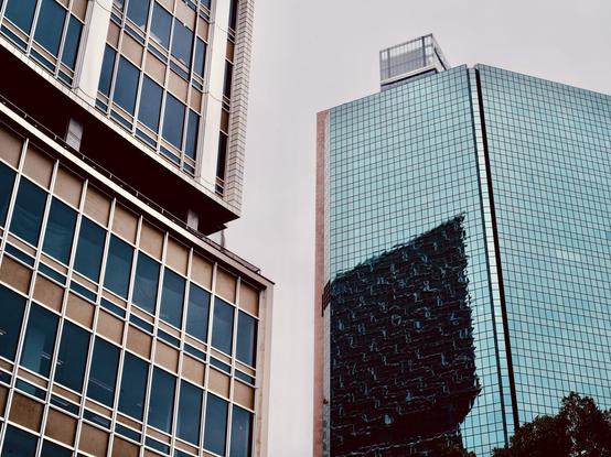 Photograph looking up at tall city buildings in Sydney.  The mid-century building on the left is a tower of steel, blue and bronze windows and white tiles. On the right is a blue glass-windowed tower. Reflected in the blue windows is a third building.
