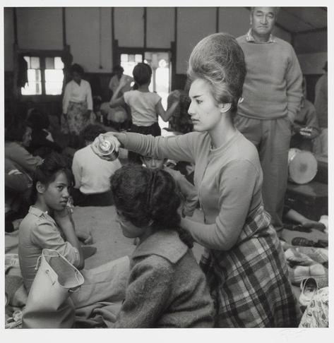 The image is a black and white photograph depicting an indoor scene with multiple people. In the foreground, there's a woman in the middle of styling another person's hair using a canister possibly containing hairspray or a similar product. The stylist has her hair styled upwards into a voluminous bouffant reminiscent of late 20th-century fashion trends.

Behind them, various individuals are seated and standing around engaged in different activities; some appear to be conversing while others seem more passive. Among the crowd is another woman who stands out due to what seems like an oversized wig or hairdo that matches the stylist's own hairstyle but appears exaggerated for comedic effect. The environment suggests a casual gathering, possibly at someone’s home during social events.

The clothing styles and hairstyles are indicative of mid-20th-century fashion trends in Japan, with traditional attire such as kimonos also visible among some individuals present. There is no explicit indication of the setting beyond that it's an indoor event where people have gathered to either celebrate or participate in a communal activity before attending church services at Ratana Pao on January 25th.

The photograph captures a slice-of-life moment, giving insight into social customs and trends from its time period.