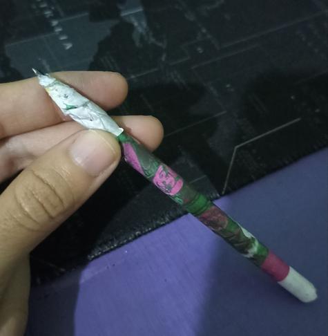 Sobre mesa morada, alguien sostiene con la mano un lápiz de papel blanco con verde y rosa, se percibe un murciélago, que es de los stickers de 1nvento. El lápiz se mira suave.
