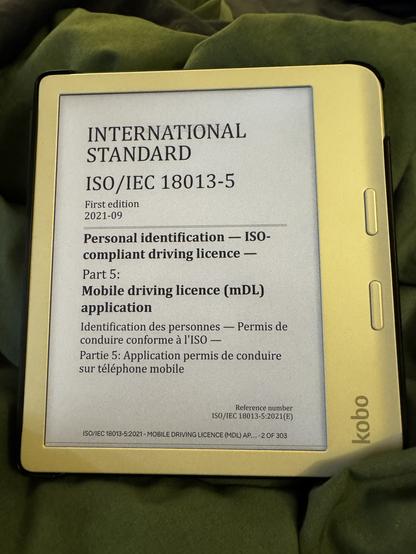 A picture of a white Kobo ereader sitting on top of a dark green comforter. The cover page of ISO/IEC 18013-5 (mobile driving license [mDL] application) is clearly visible on the ereader.