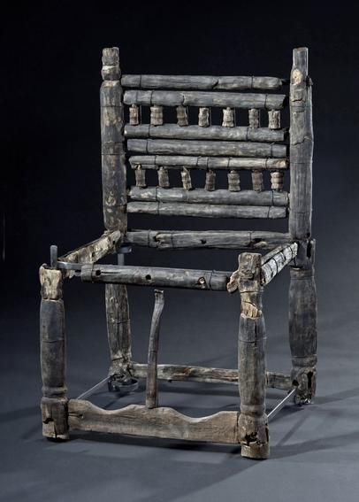 Full  shot of a wooden chair. The chair's back is a made of numerous vertical wooden slats, that are spaced evenly and have small, evenly-spaced pieces attached between each major slat. 
The chair's frame is constructed of several interconnected wooden pieces forming a rectangular base. 
The chair's seat frame is also made of a slatted design. The leather or fur seat has not survived