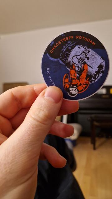 Eine Hand hält einen runden Sticker. Darauf sind ein Kosmonaut mit einer Raumkapsel. Am Rand entlang steht "Chaostreff Potsdam" und "ccc-p.org". Die Farben sind lila, orange und grau.
