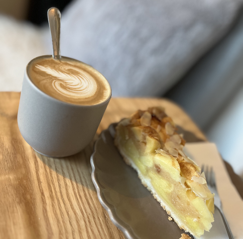 A gray cup of latte art coffee sits next to a slice of apple pie on a gray plate.  The pie has visible almond slices on top and is on a light wood table. A silver spoon rests in the coffee cup.