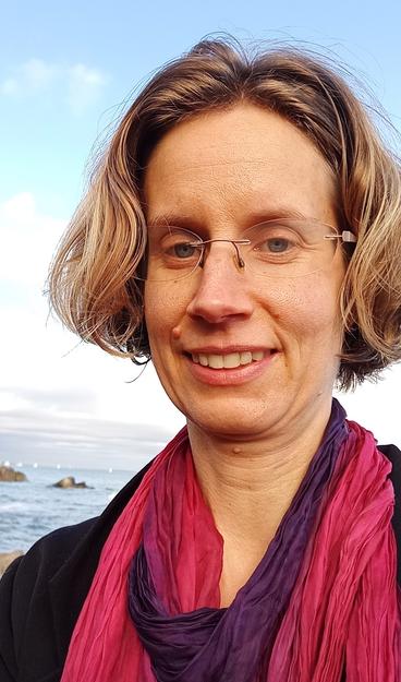 Close up of a woman with medium-length hair, wearing glasses and a pink scarf, and smiling, with a coastline and blue sky in the background