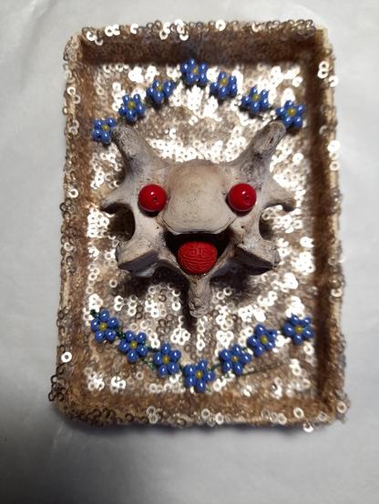 An animal vertebra looks like a smiling face from above. For emphasis, it has red glass beads as eyes and a red Asian lucky charm bead with lettering as a tongue. The monster is surrounded at the top and bottom by forget-me-nots in perlage technique - all together in a rectangular box trimmed with beige-golden sequins. Everything beams in brightness and good vibes.