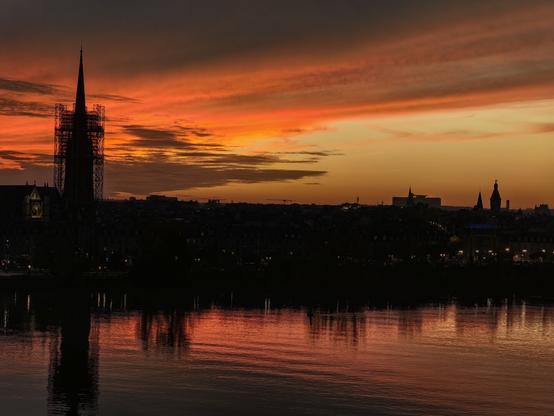 Silhouette de la ville de Bordeaux au crépuscule, avec la flèche Saint-Michel en travaux, se découpant sur un ciel flamboyant aux teintes orange et rouge, reflétées sur les eaux calmes de la Garonne.