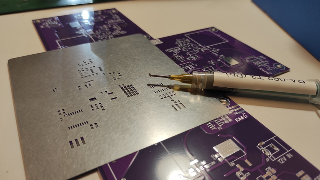 The photo shows a solder paste stencil and a syringe filled with solder paste lying atop a purple PCB. The stencil has cutouts perfectly aligned with the PCB's soldering pads, allowing precise application of paste. In the background, more PCBs are visible, hinting at a small-scale assembly