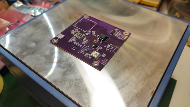 The photo displays the same purple PCB now placed on a metallic hotplate. The PCB is partially populated with small surface-mount components ready to be soldered. The reflective surface of the hotplate adds a polished, technical look to the setup.