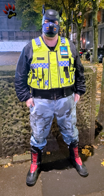 human pup, wearing camo trousers, haix boots and a hi-vis vest standing on the boardwalk in Manchester at night, full view