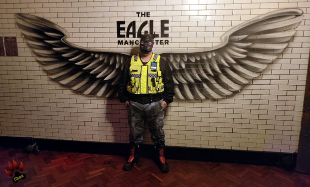 human pup, wearing camo trousers, haix boots and a hi-vis vest standing in front of the wings wall-mural at Eagle Bar, Manchester