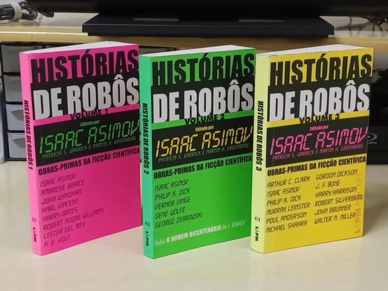 Foto de três livros de pé sobre uma mesa, cujos títulos são "Histórias de Robôs", Volumes 1, 2 e 3, respectivamente, com cores Rosa, Verde e Amarelo. No topo, o título, abaixo "Editado por Issac Asimov" em letras semelhantes à fonte de relógio digital de cristal líquido e, ao final, à esquerda, a lista de autores.
