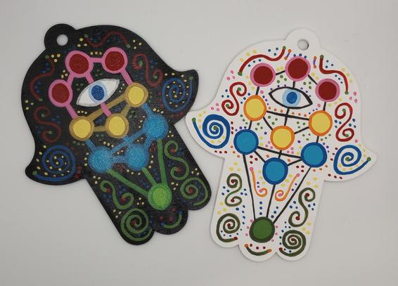 On the left a wood cutout in the shape of a hamsa hand, painted black with a colorful kabbalah tree of life design featuring an eye in the central da'at position, and glitter. 

On the right, the same wood cutout design with a white background and no glitter. 

The cutouts have a hole in the top, so they can be hung from a hook on the wall, or on a string anywhere.
