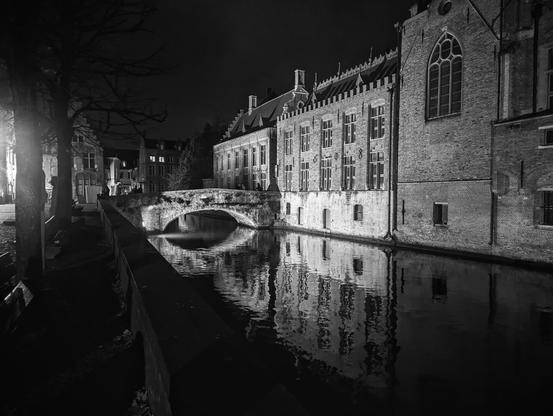 A canal, at night. There is a path and trees to the left of the canal. Buildings go right up to the water on the right hand side. There is a stone bridge in the middle distance going over the water. It's night, but the buildings  and bridge are lit up.
