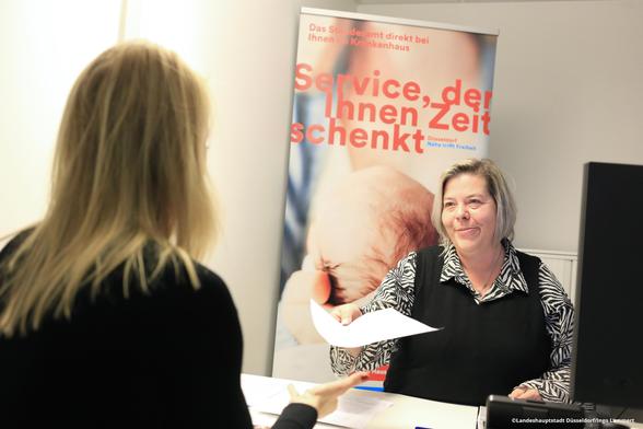 Eine Frau an einem Schreibtisch überreicht einer anderen Frau ein Blatt Papier