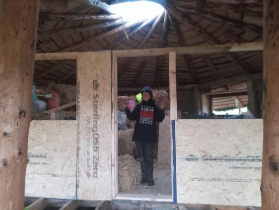Lucy stands in the door way of her new home. The front wall is very unfinished and the rest of the walls lack render, but it's now a building with a proper perimeter and a real inside and outside.