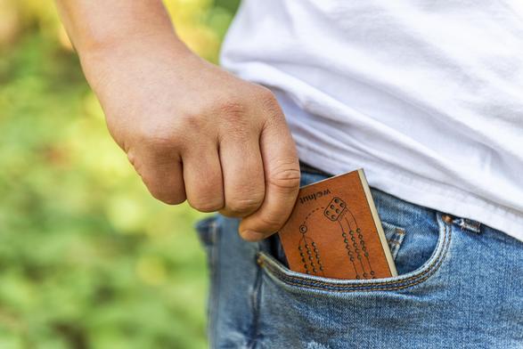 A man wearing a white t-shirt and denim jeans sliding a small leather folding cribbage board into the front pocket of jeans