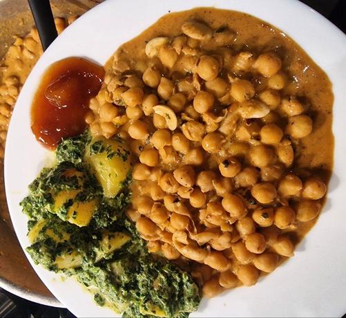 Ein weisser Teller, darauf Wetterauer Kichererbsen als Channa Masala, Aloo Palak & Chutney. Dahinter der Topf auf dem Herd mit dem Rest Kichererbsen