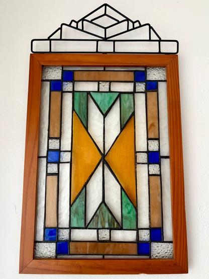 Blue, green, orange, and brown stained glass wall art in a wood frame with a metal design on top. Overall, it has an art deco look.