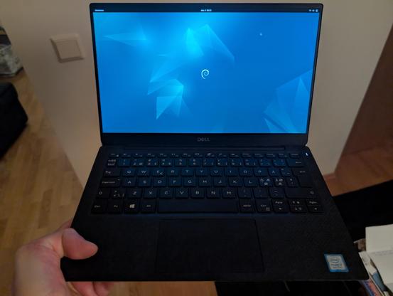 En venstrehånd som holder en åpen og kjørende Dell XPS 13 9380, med Debian Linux, hvor skjermen viser et tomt GNOME-skrivebord.

Å left hand holding an opened and running Dell XPS 13 9380, with Debian Linux, where the screen is displaying an empty GNONE desktop.