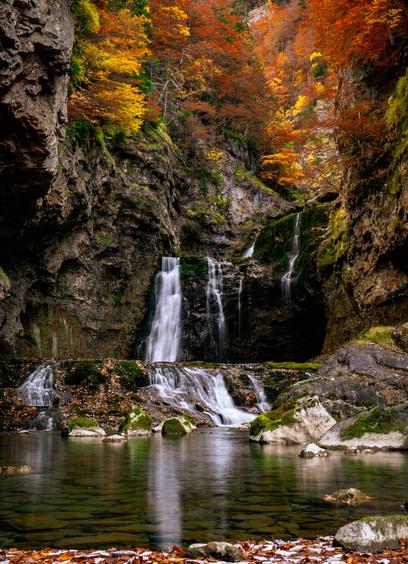 You can see a stone gorge with autumn trees in the background and a waterfall running through the gorge.