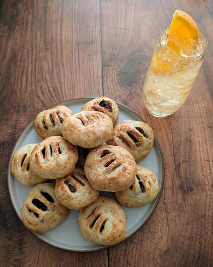 1. A photo of a plate with round puff-pastry pastries next to a cocktail, sitting on a brown wooden background. The pastries are lightly golden, with sugar sprinkled on top. Each has three slits cut in the top, through which a dark dried-fruit filling is visible. The cocktail is a light amber-coloured liquid in a high-ball glass filled with crushed ice, with an orange slice garnish.