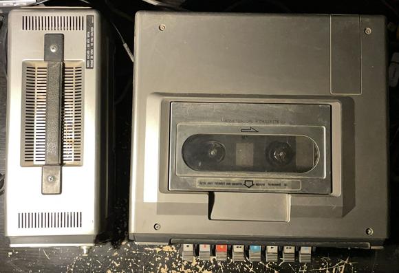 Un magnétoscope JVC HR-4100S et son alimentation externe sur un bureau vu de haut.
On voit la trappe transparente du compartiment cassette.
