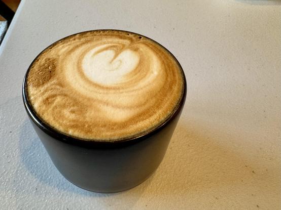 Small black coffee mug filled with a cappuccino sitting on a textured gray white table.