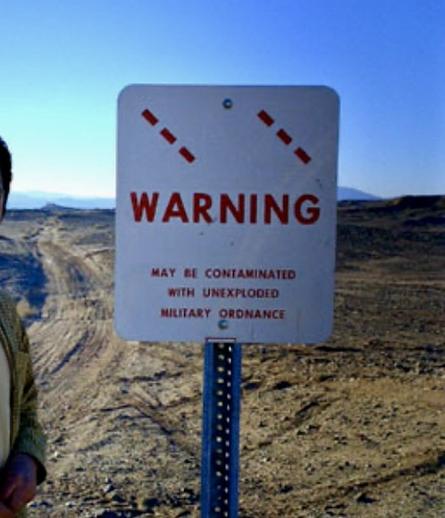 A warning sign in the middle of desert under the blue sky. It says - may be contaminated with unexploded military ordnance.