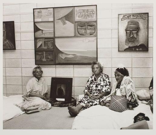 The image depicts a group of elderly women sitting on the floor in what appears to be an art gallery or exhibition space. The room has white tiled walls, and there are various artworks displayed on them, including paintings and sculptures.

Four women can be seen seated together; one is wearing a patterned black-and-white dress with glasses, while another wears a striped shawl over her shoulders. A third woman sits next to the first two, also in a traditional attire that includes a headscarf or wrap around her neck. The fourth woman appears more distanced from the group and leans against what looks like a white bench.

Behind them is an artwork titled "Capt. Bryce," which features a portrait of a man wearing a cap with text above it reading 'Capt. Bryce.' Another piece, possibly created by Te Whiti himself or someone closely associated with him, shows a black-and-white photograph framed and hung on the wall next to another abstract painting.

In front of them is an open book lying flat, suggesting that they might be engaging in conversation about art or discussing the works displayed around them. The overall atmosphere seems contemplative and serene as these women gather together within this space dedicated to artistic expression.
