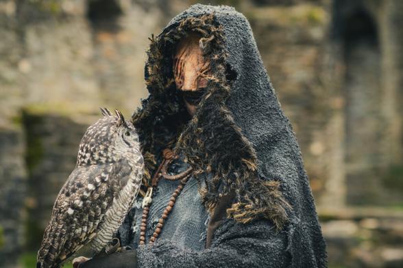 Fotografie eines Cosplayers, der den Seher aus der TV-Serie Vikings darstellt. Die Figur trägt einen dunklen, strukturierten und zerschlissenen Umhang mit einer Kapuze, die Teile des Gesichts verdeckt und eine unheimliche, mysteriöse Atmosphäre erzeugt. Das Gesicht ist mit tiefen Falten und Narben geschminkt. Der Seher hält eine Eule auf seinem Arm, im Hintergrund sind alte Steinruinen zu sehen. Schlüsselwörter: Vikings Cosplay, Der Seher, historisches Drama, düstere Fantasie, nordische Mythologie, Eulensymbolik, Wikinger-Kostüm, mittelalterliches Cosplay, Cosplay-Fotografie.