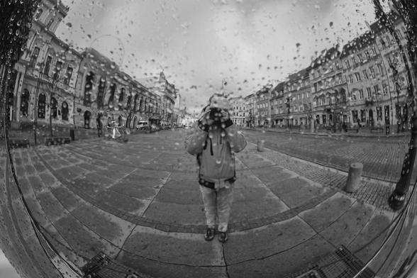 The image shows a black and white reflection of a city street scene captured in a reflective surface, possibly a spherical or curved mirror. There are buildings lining the street, and a person is taking a photo, evidenced by the camera visible in the reflection. The surface has raindrops on it, suggesting a rainy or drizzly day.
