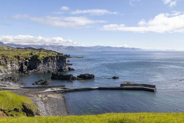 A sunny day with the edge of a chilly wind to it and we're standing on a grassy cliff top looking over the edge to a small cove with more cliffs jutting out around its far edge, a calm sea, and, on the horizon, hilly and mountainous landscape faded with distance. The cove has a shallow beach of black sand and a long stone jetty that sticks out for a hundred metres or so then turns sharply ten degrees towards us and sticks out a little further still. The cliffs of the far side of the cove are sheer and dark, suggesting this is an area formed from volcanic activity in the past, and the upper surface is covered in short, bright grass which tells us this is spring or early summer. A few thin patches of cloud are in the otherwise bright blue sky.