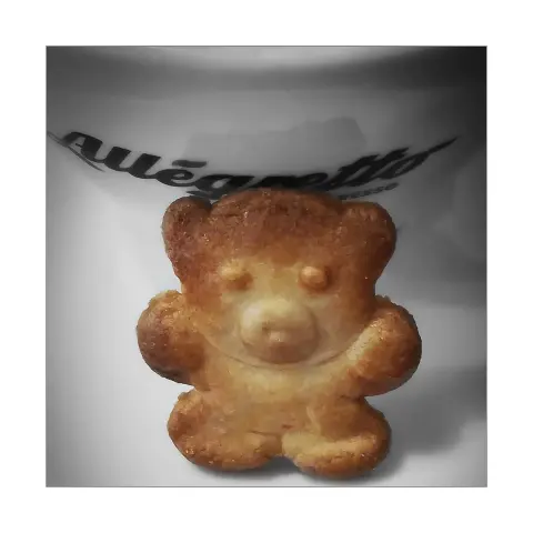 Das quadratisch beschnittene Foto zeigt im Hintergrund weißes Porzellan mit der Aufschrift 'Allegretto', darunter in kleiner Schrift 'Espresso', was auf eine Tasse oder Ähnliches weist. Daran angelehnt ein Plätzchen oder Gebäck in Form eines kleinen Bären. Ohren, Augen, Schnauze und Tatzen sind modelliert und verleihen dem Gebäck einen plastischen Eindruck .