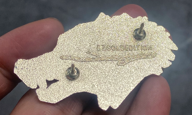 Photo of a hand holding up the reverse side of a snarling wolf PCB pin, show the pin post placement and back engraving. The engraving says "CZGOLDEDITION" above a line illustration of a stick that is half naturalistic, half printed circuit board traces.