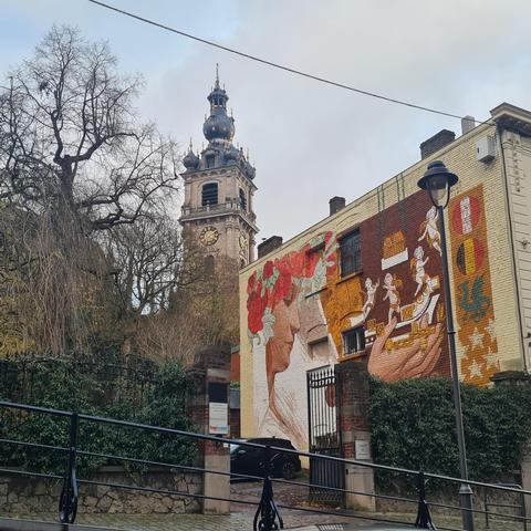A street photograph containing a cast iron gate overgrown with ivy, a building within displaying a large mural of a serene woman releasing angels from her hands. In the background an intricate church tower.