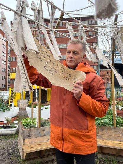 Chris Packham in an orange coat holds up a strip of linen on which he has written a message "winning is not giving up and the fuel is love and rage". Behind him are Brink's temporary planters and shipping container meeting space, surrounded by modern office and apartment blocks.
