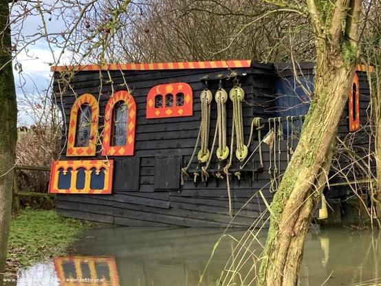 The side view of the shed, showing its dark wooden exterior with colorful, castle-like window frames and hanging ropes. The shed sits near a flooded area, and its reflection is visible in the water.