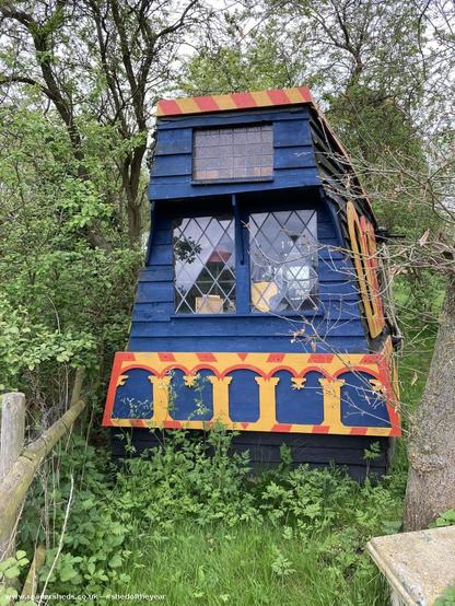 The exterior of a tall blue shed with ornate yellow and red trim, mimicking the stern of a ship. It is surrounded by trees and greenery. The shed has diamond-paned windows.