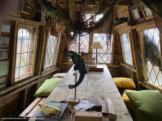 The interior of a shed, designed to resemble the captain's quarters of a ship. A long wooden table with cushions along the sides dominates the space. Diamond-paned windows provide natural light. Nets, nautical decor, and a parrot sculpture on a stand contribute to the theme.