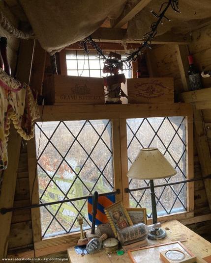 Another interior view of the shed, with a focus on a diamond-paned window. Wine boxes are used for storage on a shelf above the window. A lamp and various trinkets are on a table in the foreground