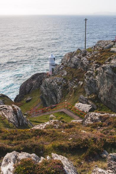 A small white lighthouse perched on a rugged coastal cliff edge, with red safety railings leading down a narrow path. The rocky cliffs are covered with wild coastal vegetation and colorful wildflowers in yellow and pink hues. The Atlantic Ocean stretches to the horizon, with choppy waters visible below. A utility pole stands on the cliff top. The scene captures the dramatic coastal landscape typical of Ireland's shoreline.