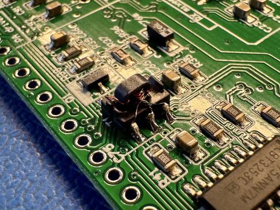 An SMD part soldered onto a green PCB. The SMD component has a ferrite ring stuck to its top. Multiple enameled wires in red and green color are wound through the ferrite ring and the wires are soldered to the legs of the SMD component. The SMD component also has lips at the end of the part to keep the ferrite ring in position.
