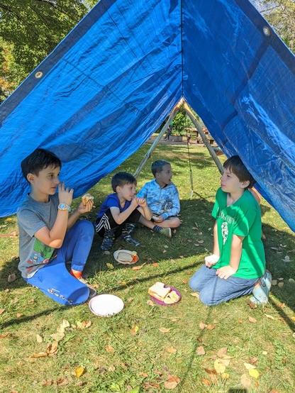 Four kids eating lunch outside on a sunny day under a tent made from a blue tarp and some sticks.