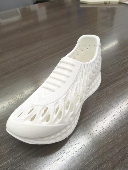 A mini tennis shoe that appears to be made of white plastic, sitting on a faux wood table. It has a strange pattern for side walls of oddly shaped organic looking sinuous holes, such that could activate your trypophobia!
