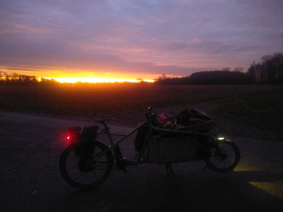 Ein Lastenrad mit gefülltem Korb steht auf einem Weg in der Dämmerung. Zwischen dem Horizont und den Wolken am Himmel gibt es eine orangerot aufgehende Sonne.