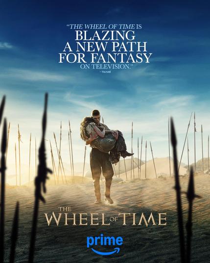 A poster of Rand al'Thor, played by Josha Stradowski, carrying Moiraine Damodred, played by Rosamund Pike, through a desert landscape surrounded by numerous spears sticking out of the sand. Text at the top reads “‘The Wheel of Time is blazing a new path for fantasy on television.’ – Vulture”. At the bottom there is The Wheel of Time and Prime logo.